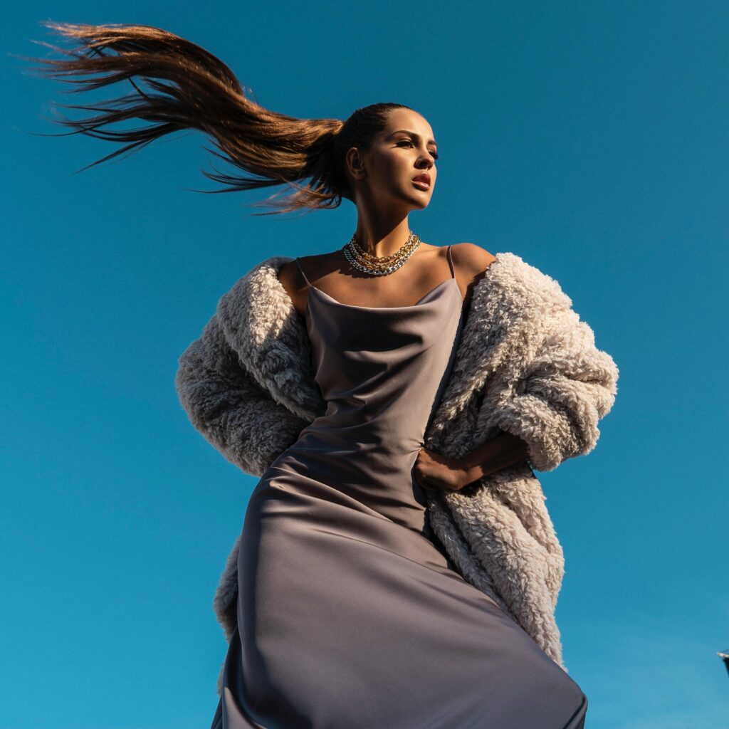 A woman in a stylish dress and fur coat poses confidently with flowing hair.